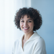 Headshot portrait of a smiling woman with short dark curly hair, wearing a white sweater, against a soft blue and white backg
